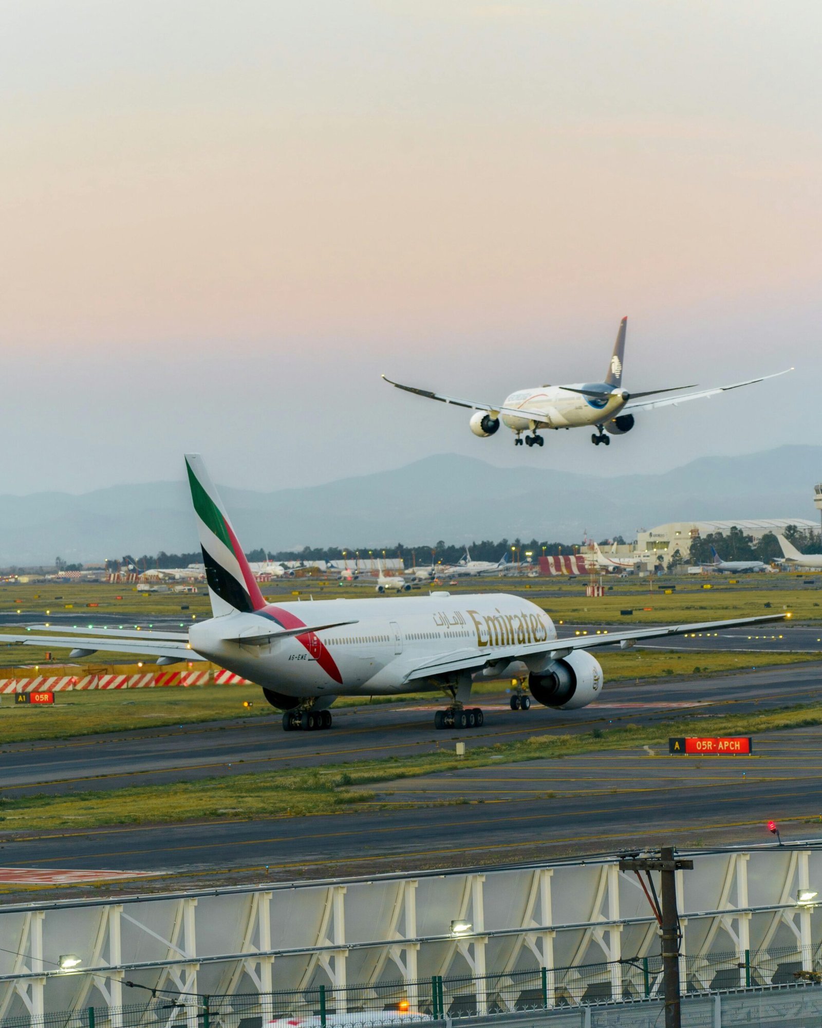 Two airplanes on the runway at sunset, one landing and one preparing for take-off, at a bustling airport.